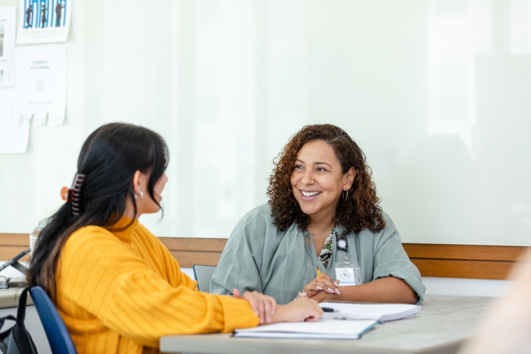 A cheerful college professor smiles as she listens to a female student discuss something during a tutoring session.